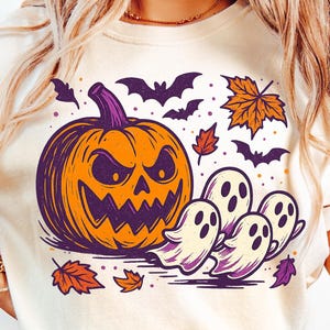 May include: Cream-colored t-shirt featuring a Halloween-themed graphic. The design includes an orange jack-o'-lantern, purple bats, and ghosts, with autumn leaves scattered around. The shirt is paired with light blue jeans.