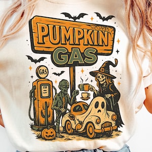 Peut inclure: T-shirt crème avec un motif sur le thème d'Halloween. Le design comprend une enseigne « Pumpkin Gas », des chauves-souris et des personnages de dessins animés à une station-service. Le t-shirt est porté avec un jean bleu clair.