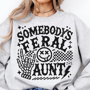 May include: A light gray sweatshirt with the text "SOMEBODY'S FERAL AUNTY" in black. The design includes a skeleton hand, a smiley face, and a checkered pattern. The sweatshirt is a casual style.