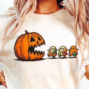 Puede incluir: Camiseta color crema con un diseño de Halloween. El diseño incluye una gran calabaza naranja con la boca abierta y tres pequeñas figuras de fantasmas. La camiseta se combina con vaqueros de mezclilla azul claro.