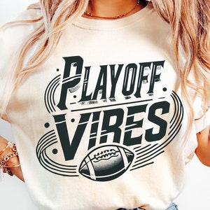 Puede incluir: Una mujer con una camiseta de color crema con el texto "PLAYOFF VIBES" y un gráfico de fútbol. También lleva pantalones vaqueros de color azul claro.