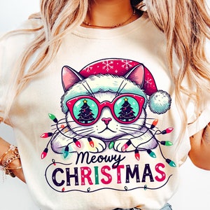 Peut inclure: T-shirt crème avec un chat de dessin animé portant des lunettes de soleil avec des lentilles en forme de sapin de Noël et un bonnet de Père Noël. Le chat est entouré de guirlandes lumineuses, avec les mots "Meowy Christmas" en couleur.
