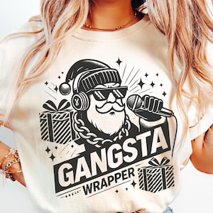 Könnte beinhalten: Ein weißes T-Shirt mit einer schwarz-weißen Illustration von Weihnachtsmann mit Kopfhörern, Sonnenbrille und einer goldenen Kette. Der Text "Gangsta Wrapper" ist unter der Illustration gedruckt.