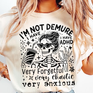 Op de afbeelding: Een wit T-shirt met een zwart-witte afbeelding van een skelet dat een beker met een rietje vasthoudt. De tekst op het shirt luidt "I'm not demure I have ADHD Very forgetful very chaotic very anxious".