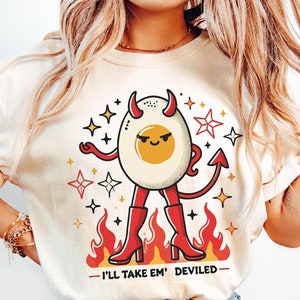 Könnte beinhalten: Ein weißes T-Shirt mit einer Cartoon-Illustration eines teuflischen Eies, das rote Stiefel und einen Teufelschwanz trägt. Das Ei hat ein gelbes Eigelb und ein rotes Gesicht. Der Text "I'll take 'em' deviled" ist unter dem Ei gedruckt.