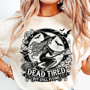 May include: A white t-shirt with a black and white graphic of a skeleton witch flying on a broomstick in front of a full moon. The text "Dead Tired But Still Flying" is written on a banner below the witch.