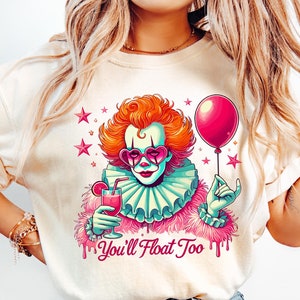 Può includere: Una maglietta bianca con una grafica di un clown che tiene un palloncino rosa e un bicchiere di liquido rosso. Il clown indossa occhiali da sole a forma di cuore rosa e un colletto a volant rosa. Il testo "You'll Float Too" è stampato sotto la grafica.