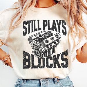 Peut inclure: Une femme portant un t-shirt beige clair avec un graphique noir et blanc d'un moteur et le texte "Still Plays Blocks". Elle porte un jean bleu clair.
