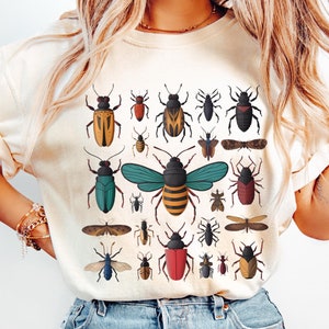 May include: A beige t-shirt with a graphic print of various insects in different colors, including red, blue, green, yellow, and brown.