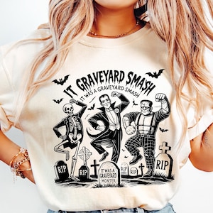 May include: A white t-shirt with a black and white graphic print of a skeleton, a man in a tuxedo, and a monster dancing in a graveyard. The text on the shirt reads "It Graveyard Smash It Was A Graveyard Smash".