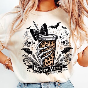 May include: A white t-shirt featuring a graphic of a skeleton hand holding a leopard print coffee cup with a black bow on top. The graphic is surrounded by bats, skulls, and stars. The text "BOO-jee Mama" is written on a black ribbon below the graphic.