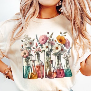 Può includere: Una maglietta bianca con un design floreale con fiori colorati in becher di vetro. I fiori sono rosa, bianchi, gialli e arancioni.