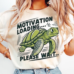 Può includere: Una maglietta beige con una tartaruga verde dei cartoni animati e il testo "Motivation Loading... Please Wait..."