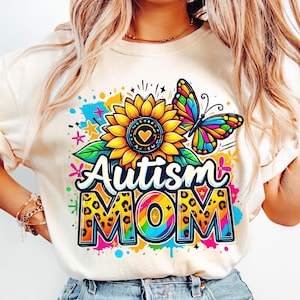 May include: A white t-shirt with a colorful graphic design featuring a sunflower, a butterfly, and the text "Autism Mom" in a rainbow and leopard print design.