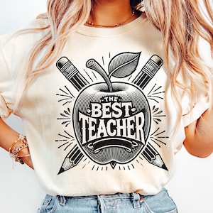 May include: A white t-shirt with a black graphic of an apple with the text "The Best Teacher" inside the apple. Two pencils are crossed behind the apple.