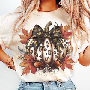 Puede incluir: Una camiseta beige con un gráfico de una calabaza de estampado de leopardo con un lazo marrón y dorado. La calabaza está rodeada de hojas de otoño en tonos naranja, rojo y amarillo.