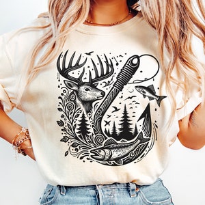 Op de afbeelding: Een crèmekleurig T-shirt met een zwart grafisch ontwerp van een hert, een vis en een haak met een bos scène op de achtergrond.