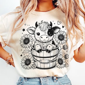 May include: A cream colored t-shirt with a black and white illustration of a cow in a bucket with sunflowers.