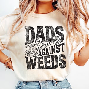 Op de afbeelding: Een wit T-shirt met een zwarte afbeelding waarop "Dads Against Weeds" staat met een illustratie van een grasmaaier.