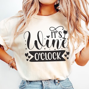 May include: A woman wearing a light beige t-shirt with the text "It's Wine O'Clock" in black lettering and a wine glass icon. She is also wearing light blue jeans.