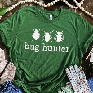 May include: Green t-shirt with a white graphic of three bugs and the text "bug hunter". The shirt is laid out on a wooden surface with a pair of gardening gloves, a watch, a pair of jeans, and a plant.