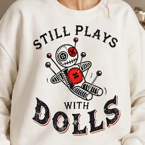 May include: White sweatshirt with a black graphic of a voodoo doll with the text "Still Plays With Dolls" printed on it.