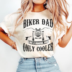 Könnte beinhalten: Eine Frau trägt ein hellbeiges T-Shirt mit dem Text "Biker Dad Like a Normal Dad Only Cooler" und einer schwarzen Grafik eines Motorrads. Sie trägt auch hellblaue Jeans.