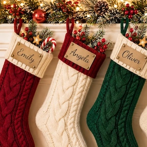 May include: Four cable-knit Christmas stockings hanging in a row. The stockings are red, cream, green, and cream, each featuring a cream-colored cuff with a rectangular leather tag engraved with the names Emily, Angela, Oliver, and Mason.