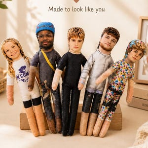 May include: Five custom fabric dolls featuring printed photographic faces and various outfits, including a white shirt, a dark jumpsuit with a yellow strap, a black outfit, a light gray shirt, and a colorful floral patterned shirt with a hood.