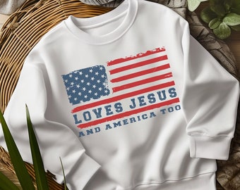 Może przedstawiać: Biała bluza z nadrukiem przetartej flagi amerykańskiej i tekstem "Loves Jesus and America Too".