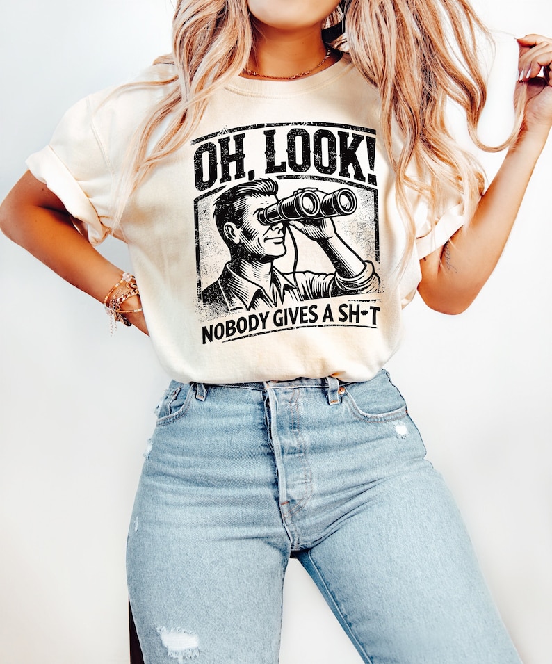 May include: A cream colored t-shirt with a black graphic of a man looking through binoculars. The graphic has the text "Oh, Look! Nobody Gives A Sh*t" underneath it.