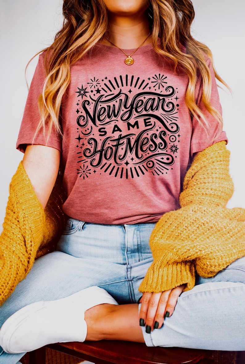 May include: A woman wearing a pink t-shirt with the text "New Year Same Hot Mess" in black lettering. The shirt has a festive design with fireworks and stars.