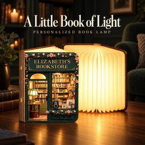 May include: A book-shaped lamp featuring a miniature bookstore diorama titled Elizabeth's Bookstore on the front cover, decorated with pink flowers. The lamp is open, displaying a warm, illuminated interior with shelves of books and a small table lamp.
