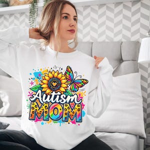 May include: White sweatshirt with a colorful graphic design featuring a sunflower, butterfly, and the text "Autism Mom".