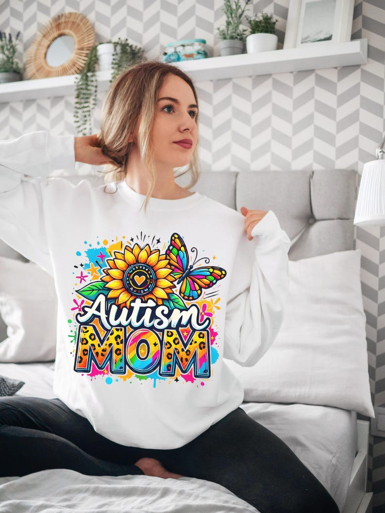 May include: White sweatshirt with a colorful graphic design featuring a sunflower, butterfly, and the text "Autism Mom".