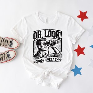 May include: White t-shirt with a black and white illustration of a person looking through binoculars. The text "OH LOOK! NOBODY GIVES A SH-T" is printed on the shirt.