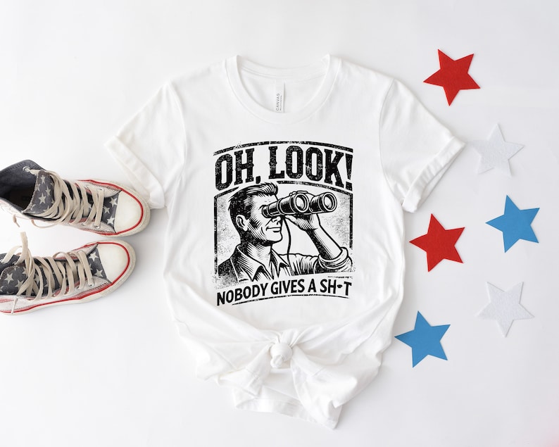 May include: White t-shirt with a black and white illustration of a person looking through binoculars. The text "OH LOOK! NOBODY GIVES A SH-T" is printed on the shirt.