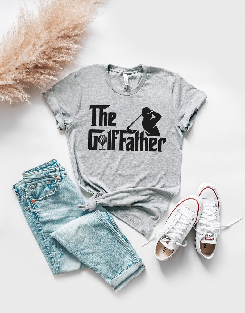 May include: A light grey t-shirt with the text "The GolfFather" in black, featuring a golf ball and golfer silhouette. The shirt is paired with light blue jeans and white trainers with red accents. A casual, sporty theme.