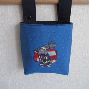 Könnte beinhalten: Blauer Stoffbeutel mit strukturierter Oberfläche und marineblauen Besätzen und Trägern. Die Tasche zeigt ein gesticktes Bild eines Elefantenfeuerwehrmanns vor einem roten Feuerwehrauto. Die Tasche hängt an einer Holzstange.