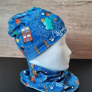 Könnte beinhalten: Blaues Mütze- und Halstuch-Set mit London-Motiv. Der Stoff zeigt Illustrationen eines roten Doppeldeckerbusses, Sehenswürdigkeiten und die Schriftzüge "Windsor Gardens" und "Paddington". Das Set wird auf einem Schaufensterkopf präsentiert.