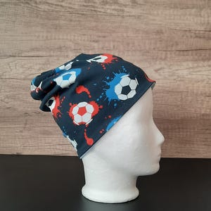 Könnte beinhalten: Eine marineblaue Beanie-Mütze mit einem Fußballmuster und roten, weißen und blauen Farbspritzern. Die Mütze wird auf einem weißen Schaufensterkopf gezeigt. Die Mütze ist aus weichem Material.