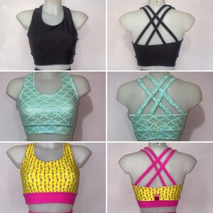 Op de afbeelding: Drie verschillende sportbeha's met verschillende patronen en rugbandjes. De eerste beha is zwart met een racerback-stijl. De tweede beha is teal met een zeemeermin-schubbenpatroon en gekruiste bandjes. De derde beha is geel met een bliksem-patroon en gekruiste bandjes.