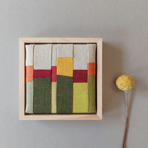 Könnte beinhalten: Ein quadratisches Textilkunstwerk mit vertikalen Stoffstreifen in Olivgrün, Limettengrün, Gelb, Rot, Orange und Beige, eingefasst in einen hellen Holzrahmen. Ein einzelner getrockneter gelber Blumenstängel liegt auf der rechten Seite.