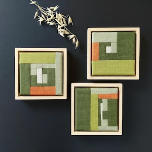 Könnte beinhalten: Drei quadratische Wandkunstwerke mit abstrakten geometrischen Designs. Jedes Stück zeigt ein Patchwork aus grünen, orangefarbenen und grauen Stoffquadraten in einem Naturholzrahmen. Ein Zweig getrockneter Blumen befindet sich in der oberen linken Ecke.