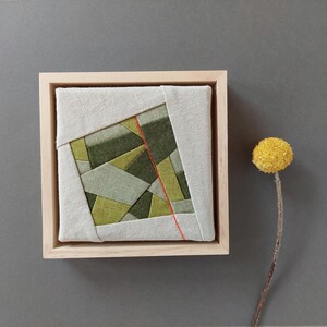 Könnte beinhalten: Gerahmtes Textilkunstwerk mit geometrischem Muster in Grüntönen, akzentuiert durch eine dünne rote Linie. Das Werk ist in einem hellen Holzrahmen gefasst. Eine getrocknete gelbe Blume befindet sich auf grauem Hintergrund.