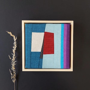 Könnte beinhalten: Abstrakte Textilkunst in einem hellen Holzrahmen. Das Kunstwerk zeigt ein geometrisches Design mit Farbblöcken, darunter Türkis, Rot, Hellblau, Lila und Weiß. Das Kunstwerk ist auf einem dunklen Hintergrund montiert.