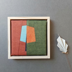 Könnte beinhalten: Ein quadratisches Textilkunstwerk in einem hellen Holzrahmen mit geometrischen Stoffbahnen in Rostrot, Olivgrün, leuchtendem Orange und Hellblau. Neben dem Rahmen liegt ein kleines, weißes, gefaltetes papierartiges Fächerobjekt mit Stiel.