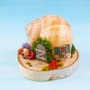 Könnte beinhalten: Ein Miniatur-Schneckenhaus mit einer roten Tür und einem Fenster, umgeben von Grün und Blumen, auf einem Holzsockel. Zwei Miniaturfiguren sitzen auf einer roten Bank.