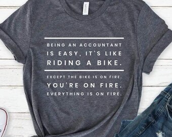 May include: A dark heather gray short-sleeve t-shirt featuring white text that reads: BEING AN ACCOUNTANT IS EASY, IT'S LIKE RIDING A BIKE. EXCEPT THE BIKE IS ON FIRE. YOU'RE ON FIRE. EVERYTHING IS ON FIRE. The shirt is paired with blue denim jeans.