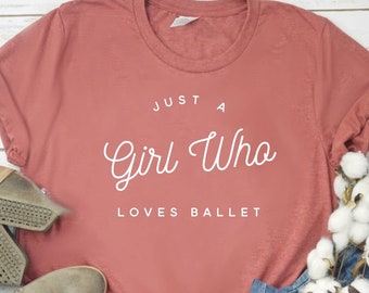 May include: A dusty rose short-sleeve t-shirt with white text that reads JUST A Girl Who LOVES BALLET, paired with blue denim jeans and taupe perforated ankle boots, accented by a decorative cotton stem and a small fabric pumpkin.